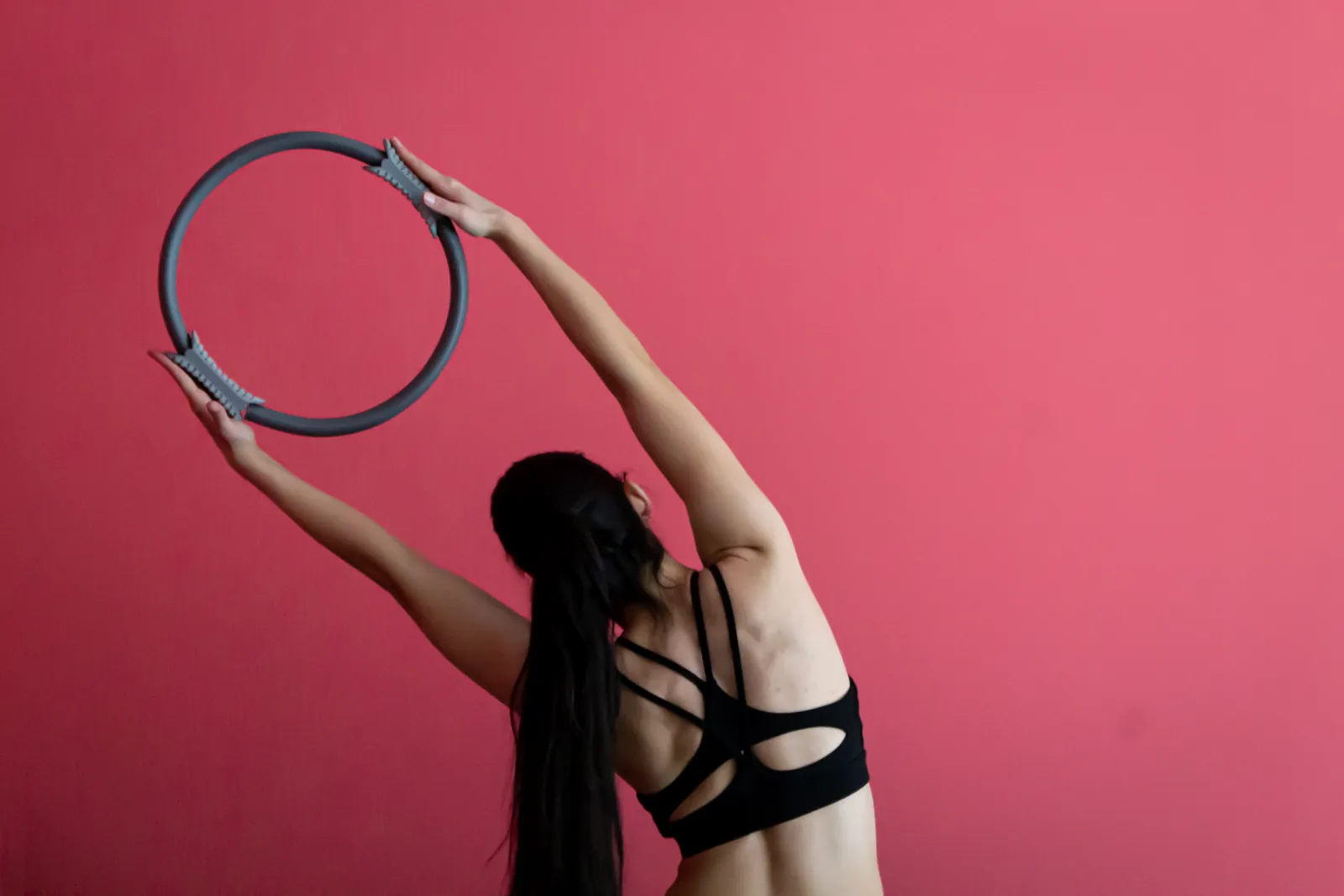 De espaldas, una mujer sostiene el aro de Pilates sobre la cabeza frente al muro rosado del estudio.