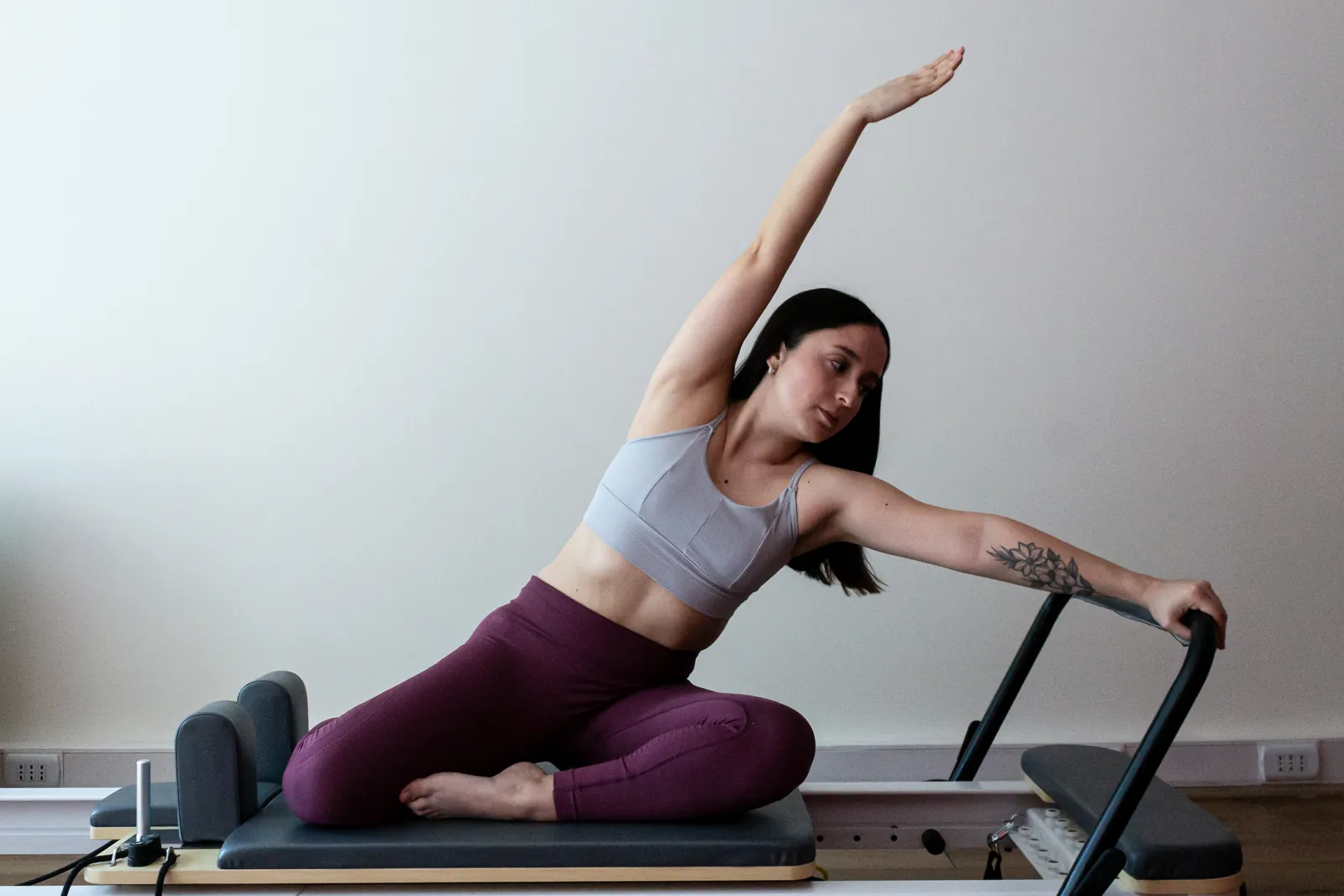 Mujer sentada sobre el reformer en un estiramiento lateral, con un brazo en alto y la otra mano en la barra de la máquina.